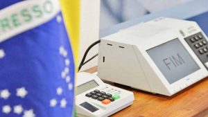 Urna eletrônica em cima de uma mesa ao lado da bandeira do Brasil